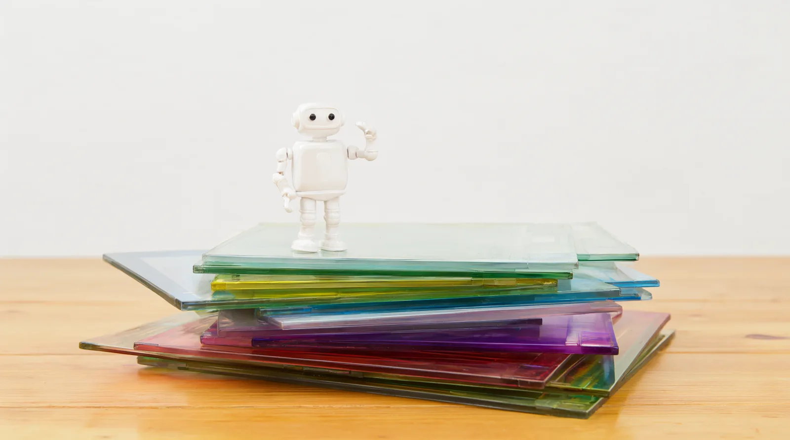 Kleine weiße Roboterfigur steht auf einem Stapel bunter, durchscheinender Glasplatten auf einem hellen Holztisch