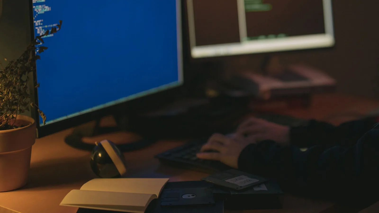 Eine Person tippt im abgedunkelten Raum an einem Arbeitsplatz mit zwei Monitoren, auf denen Quellcode zu sehen ist