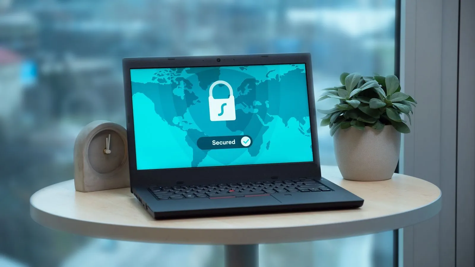 Aufgeklappter Laptop mit einem Schloss-Symbol auf dem Bildschirm, daneben eine Topfpflanze und eine Uhr