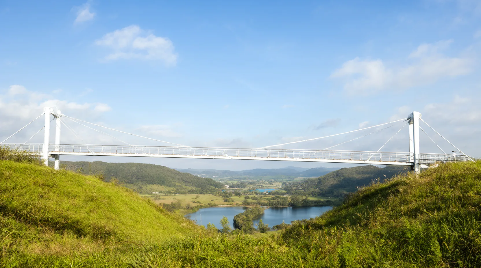 Eine schlanke weiße Hängebrücke verbindet zwei grüne Hügel über einem Tal mit einem kleinen See, darüber blauer Himmel mit leichten Wolken