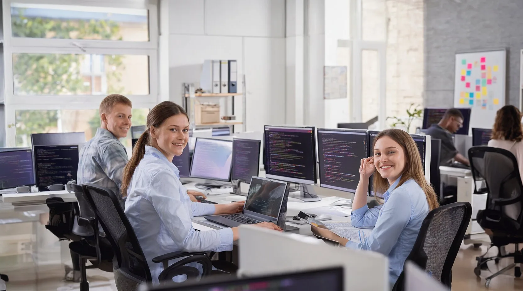 Lächelnde Softwareentwickler an Arbeitsplätzen mit mehreren Monitoren in einem hellen Großraumbüro