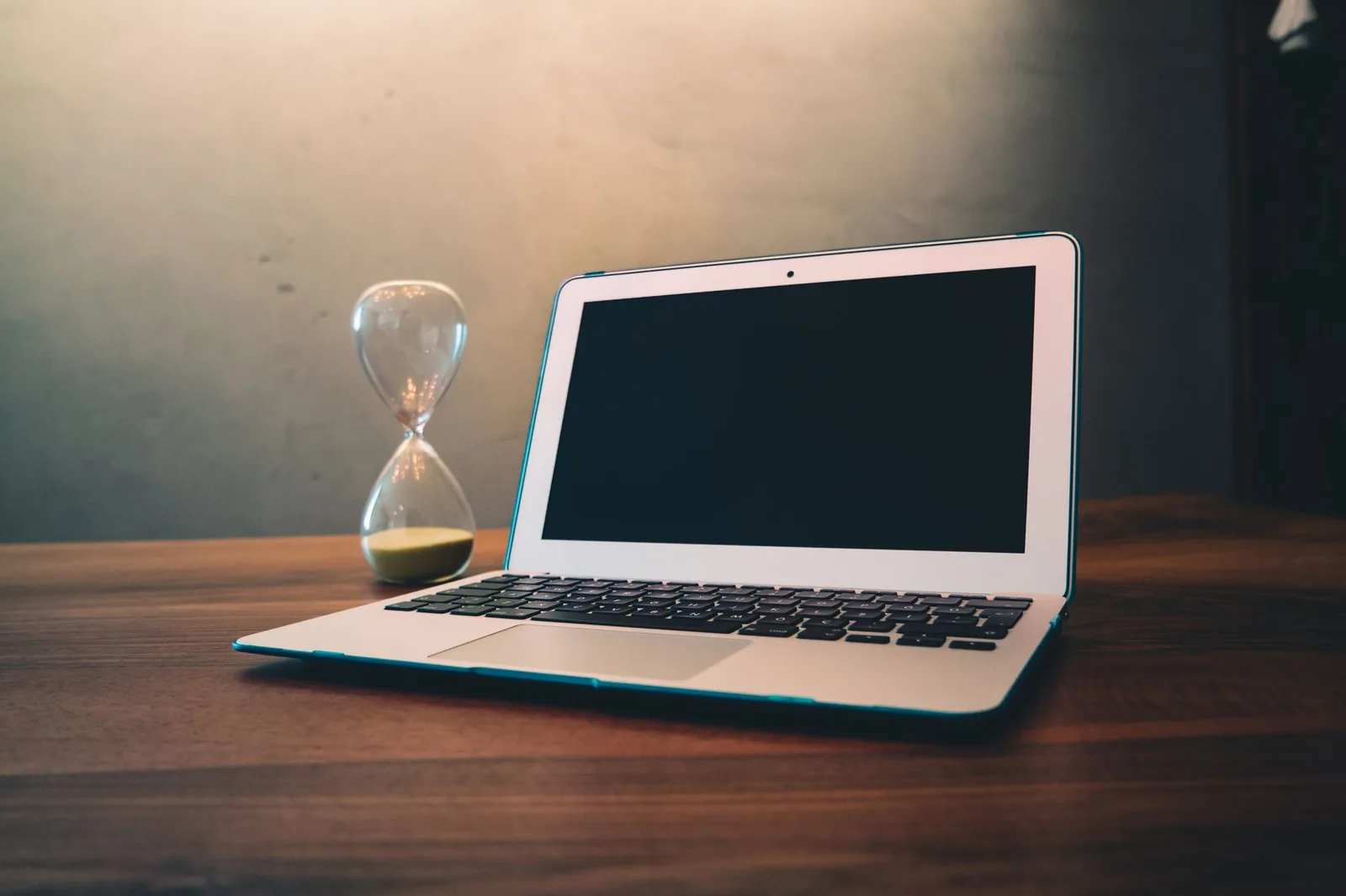 Ein aufgeklappter Laptop steht auf einem Holztisch, daneben eine Sanduhr, deren Sand zum größten Teil durchgelaufen ist