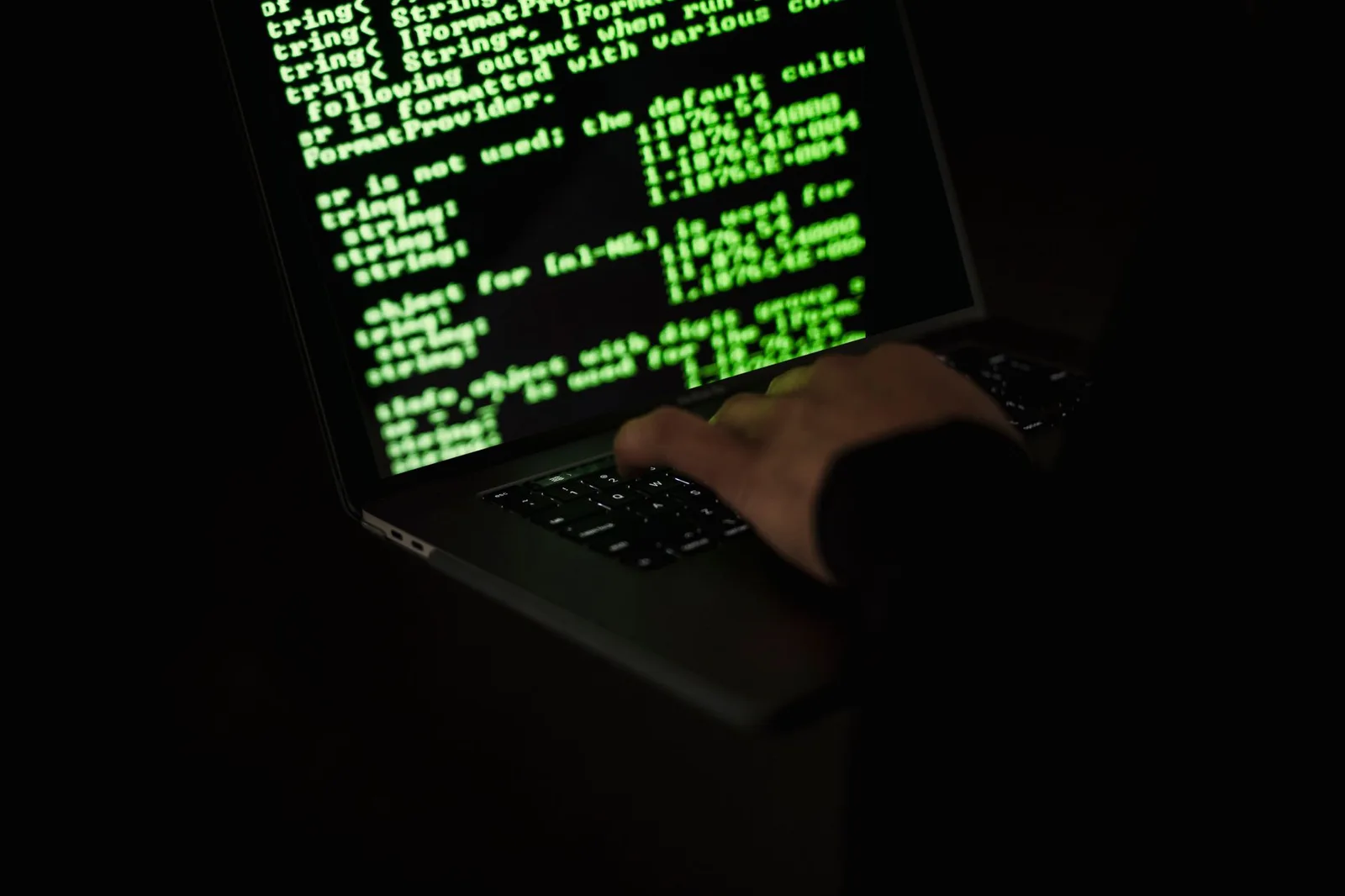 Person tippt im Dunkeln auf einem Laptop, auf dem Bildschirm Programmcode