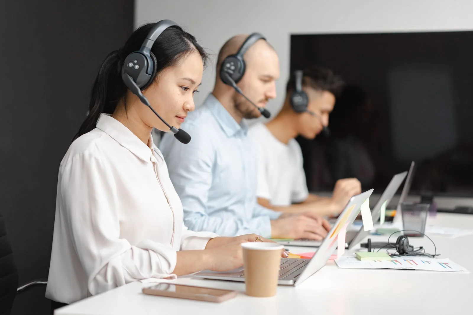 Kundenservice-Mitarbeiterin mit Headset am Laptop, dahinter zwei Kollegen, ebenfalls mit Headset, in einem hellen Callcenter